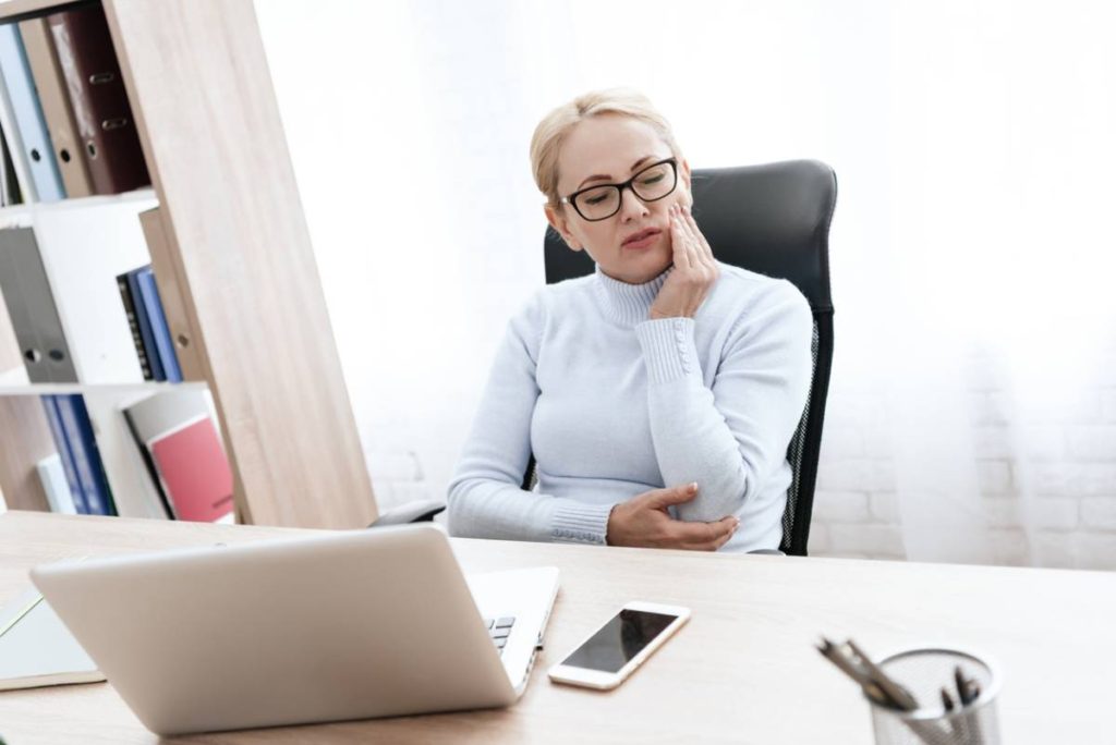 Businesswoman sitting at a desk with hand to face because of uncomfortable dentures.